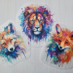 Könnte beinhalten: Drei Aquarell-Tierillustrationen auf weißem Papier. Ein Löwe mit einer rosafarbenen Mähne, ein roter Fuchs und ein Fuchs mit orangefarbenem Fell. Die Tiere sind von bunten Aquarellspritzern umgeben.