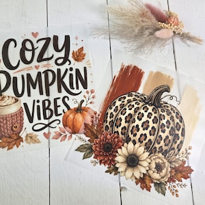 Könnte beinhalten: Zwei rechteckige Transferfolien mit herbstlichen Motiven. Ein Bogen zeigt den Text Cozy Pumpkin Vibes mit einer Tasse in einem Stricküberzug und einem Kürbis. Der zweite Bogen zeigt einen großen Kürbis mit Leopardenmuster und Herbstblumen.