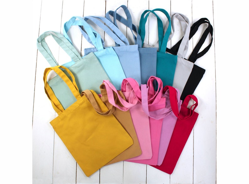 May include: A collection of 12 tote bags in various colors, including yellow, blue, green, pink, and red. The bags are made of a soft, canvas-like material and have a simple design.