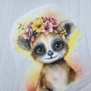 Könnte beinhalten: Eine Aquarell-Illustration eines Lemurs, der eine Blumenkrone mit rosa und gelben Blumen trägt. Der Lemur hat große braune Augen und einen süßen Ausdruck.