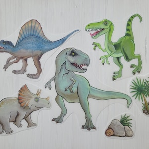 Könnte beinhalten: Fünf bunte Dinosaurier-Aufkleber, darunter ein grüner Tyrannosaurus Rex, ein grüner Velociraptor, ein blauer Spinosaurus, ein grauer Triceratops und eine grüne Palme.
