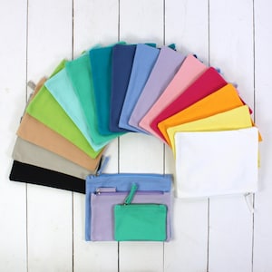 Könnte beinhalten: Eine Sammlung farbenfroher Reißverschlusstaschen in verschiedenen Farbtönen, angeordnet in einem Halbkreis. Zu den Farben gehören Schwarz, Beige, Grün, Blau, Rosa und Gelb. Im Vordergrund befinden sich drei Taschen, mit einer hellblauen, einer lila und einer grünen Tasche.