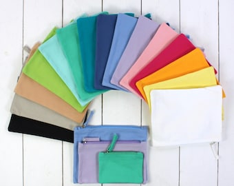 Op de afbeelding: Een verzameling kleurrijke etuis met ritssluiting in verschillende tinten, gerangschikt in een halve cirkel. Kleuren zijn onder meer zwart, beige, groen, blauw, roze en geel. Drie etuis staan op de voorgrond, met een lichtblauw etui, een paars etui en een groen etui.