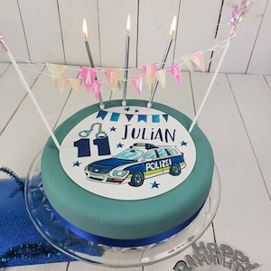 Könnte beinhalten: Eine blaue und weiße Geburtstagstorte mit einem Polizeiauto-Design und dem Namen "Julian" darauf. Die Torte ist mit einem blauen Band verziert und hat Kerzen oben drauf.