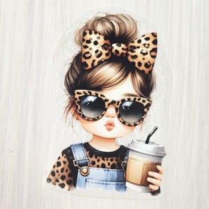 Könnte beinhalten: Illustration eines Mädchens mit braunen Haaren im Dutt, das eine Sonnenbrille und eine passende Schleife mit Leopardenmuster trägt. Sie trägt ein Oberteil mit Leopardenmuster und eine Jeans-Latzhose und hält eine Kaffeetasse mit einem schwarzen Strohhalm.