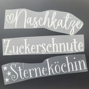 Könnte beinhalten: Drei weiße Vinyl-Aufkleber mit deutschen Wörtern auf schwarzem Hintergrund. Der obere Aufkleber sagt "Naschkatze", der mittlere Aufkleber sagt "Zuckerschnute" und der untere Aufkleber sagt "Sterneköchin".