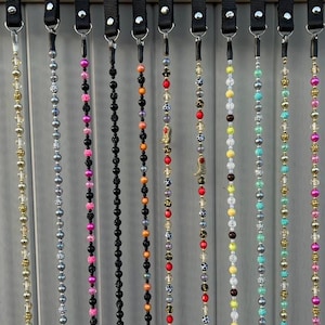 Puede incluir: Una colección de 14 cadenas de cuentas con varios colores y patrones. Cada cadena está unida a una correa negra con un clip de metal.
