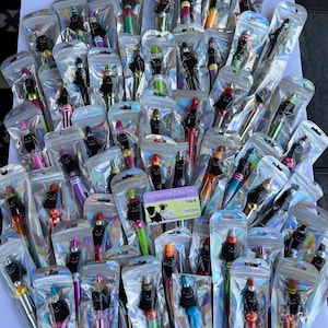 Könnte beinhalten: Eine große Sammlung von schwarz-weißen, hundeartigen Stiften in durchsichtigen Plastikverpackungen. Jeder Stift hat ein anderes farbiges Design und ist mit Perlen und anderen Verzierungen verziert. Die Stifte sind in Reihen auf einer weißen Oberfläche angeordnet. Der Text "Accessorize Your Pup" und "Ball Time" ist auf einer kleinen Karte in der Mitte des Bildes sichtbar.