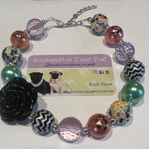 May include: Halloween-themed beaded necklace for pets, featuring black and white chevron, green, pink, orange, and black beads with a black rose charm. The necklace is on a white surface with a business card that reads "Accessorize Your Pug" and "Beaded necklaces for pets".