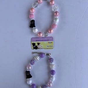 Pode incluir: Dois colares de contas com tema de cachorro. O primeiro colar é rosa com detalhes brancos e pretos. O segundo colar é roxo com detalhes brancos e pretos. Ambos os colares têm um pingente de pug preto.