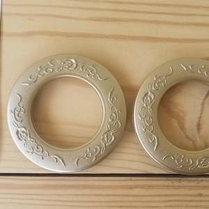 May include: Two gold-colored metal curtain rings with an ornate floral design. The rings are approximately 2 inches in diameter.