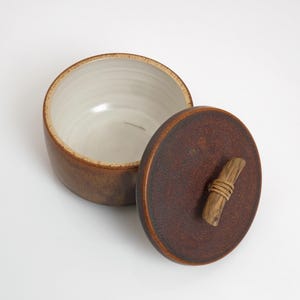 Könnte beinhalten: Ein brauner Keramiktopf mit Deckel. Der runde Topf hat einen hellen Innenraum und eine dunkelbraune Außenseite. Der Deckel ist dunkler braun mit einem Holzgriff, der mit Bindfaden umwickelt ist. Der Deckel ist leicht geöffnet.