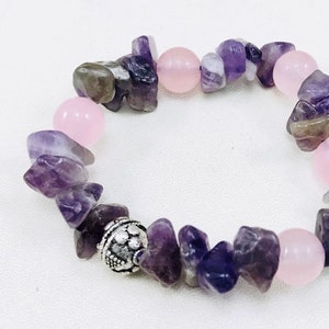 Puede incluir: Un brazalete de piedras preciosas moradas y rosas con un dije de plata. El brazalete está hecho de piedras de forma irregular y cuentas redondas.