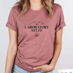 Könnte beinhalten: Ein T-Shirt in Heide-Mauve mit dem Text "Live Love Laboratory Stuff" in schwarzer Schrift. Ein kleines Herzsymbol befindet sich über dem Wort "Love".