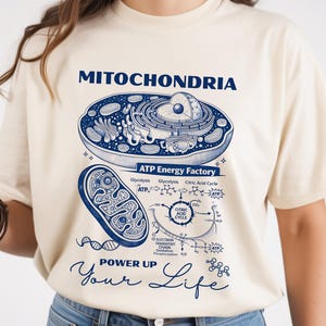 Könnte beinhalten: Ein cremefarbenes T-Shirt mit einer detaillierten blauen Illustration von Mitochondrien, beschriftet mit "MITOCHONDRIA" und "ATP Energy Factory". Darunter steht der Text "POWER UP Your Life". Ein lässiges, wissenschaftliches Kleidungsstück.