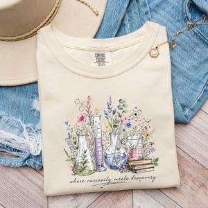 Könnte beinhalten: Cremefarbenes Comfort Colors T-Shirt mit einem Aquarelldesign von Laborgeräten und Blumen. Die Grafik enthält den Text "where curiosity meets discovery". Das Shirt wird mit Jeansshorts und einem Strohhut kombiniert.