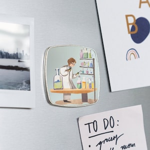 Puede incluir: Un imán cuadrado con una ilustración de dibujos animados de una persona con bata de laboratorio trabajando con botellas y vasos de precipitados de colores. El imán tiene un borde plateado.