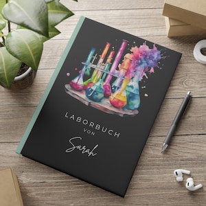 May include: A black notebook with a watercolor illustration of colorful beakers and a splash of color. The text "LABORBUCH VON Sarah" is printed on the front cover.