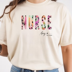 Könnte beinhalten: Cremefarbenes T-Shirt mit dem Wort "NURSE" in bunten, floral gemusterten Buchstaben. Der Name "Amy" und "Est. 2026" sind darunter gedruckt. Das Shirt ist ein lässiger Stil mit kurzen Ärmeln.