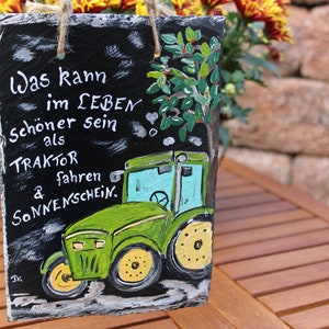 May include: A rectangular slate sign with a painted green tractor and the German text "Was kann im LEBEN sch&ouml;ner sein als TRAKTOR fahren & Sonnenschein." The sign has a jute rope for hanging.
