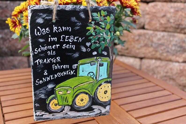 May include: A rectangular slate sign with a painted green tractor and the German text "Was kann im LEBEN sch&ouml;ner sein als TRAKTOR fahren & Sonnenschein." The sign has a jute rope for hanging.