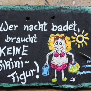 May include: Hand-painted slate sign with the German text "Wer nackt badet, braucht KEINE Bikini-Figur!" A cartoon figure in a bikini is depicted, along with a sun and two buckets. The sign has a green border.