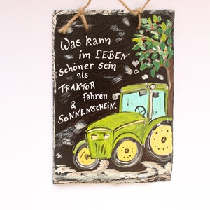 May include: Hand-painted slate sign featuring a green tractor illustration with yellow wheels and the German text "Was kann im LEBEN sch&ouml;ner sein als TRAKTOR fahren & SONNENSCHEIN." The sign is hung with a natural twine.
