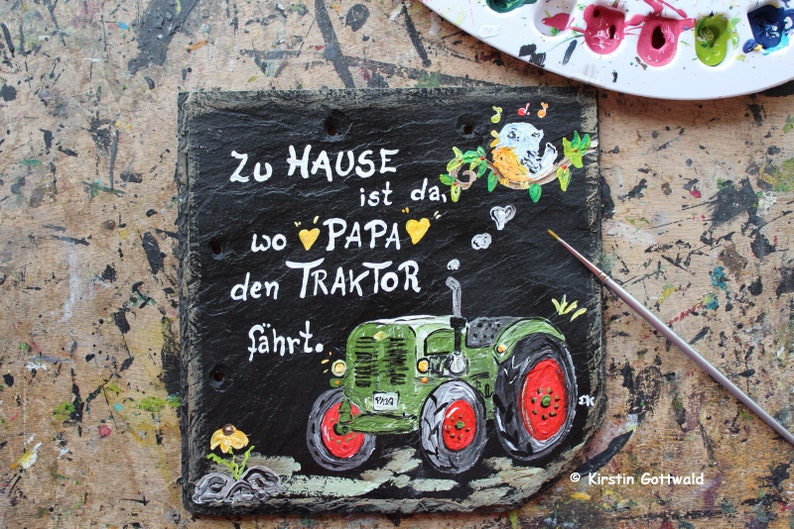 Puede incluir: Letrero de pizarra negra pintado a mano con el texto alem&aacute;n "Zu Hause ist da, wo Papa den Traktor f&auml;hrt." Presenta un tractor verde con ruedas rojas y una ilustraci&oacute;n de un p&aacute;jaro. Incluye la firma del artista.