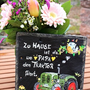Puede incluir: Un letrero de pizarra pintado a mano con el texto alem&aacute;n "Zu Hause ist da, wo Papa den Traktor f&auml;hrt" (El hogar es donde pap&aacute; conduce el tractor). El letrero presenta una ilustraci&oacute;n de tractor verde y un ramo de flores.