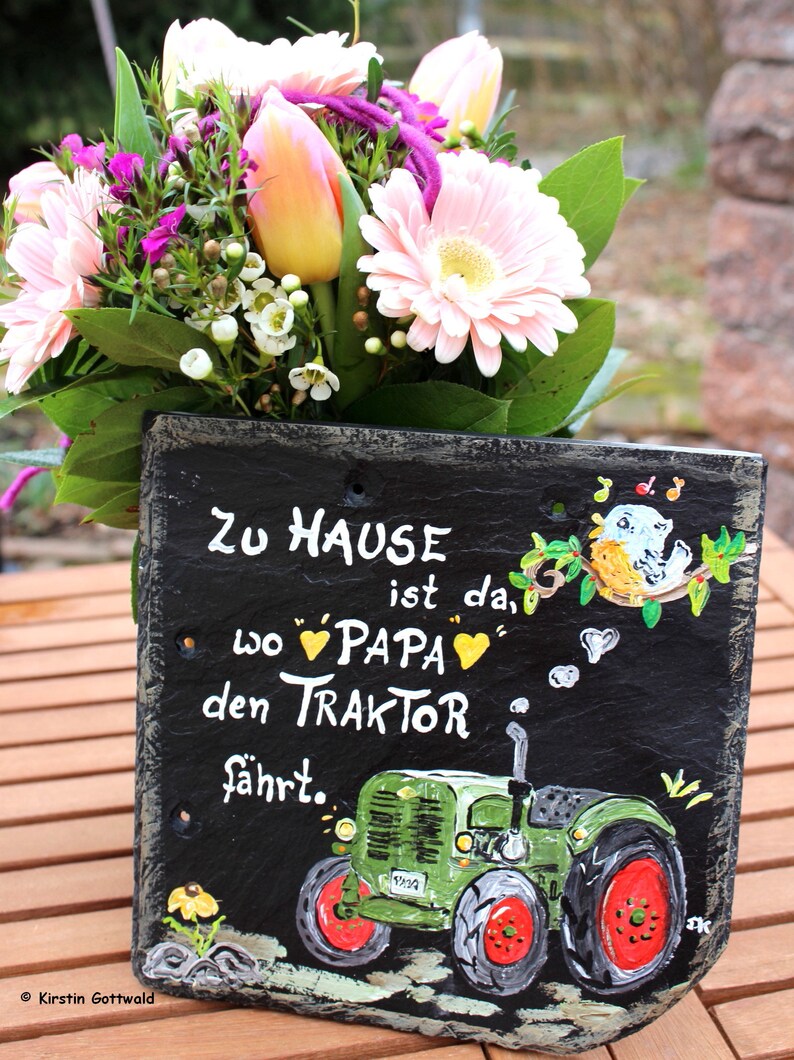 Puede incluir: Un letrero de pizarra pintado a mano con el texto alem&aacute;n "Zu Hause ist da, wo Papa den Traktor f&auml;hrt" (El hogar es donde pap&aacute; conduce el tractor). El letrero presenta una ilustraci&oacute;n de tractor verde y un ramo de flores.