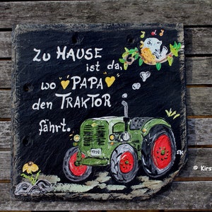 Puede incluir: Letrero de pizarra pintado a mano con un tractor verde y el texto alem&aacute;n "Zu Hause ist da, wo Papa den Traktor f&auml;hrt." Incluye detalles de p&aacute;jaros y flores. El letrero se muestra sobre una superficie de madera.