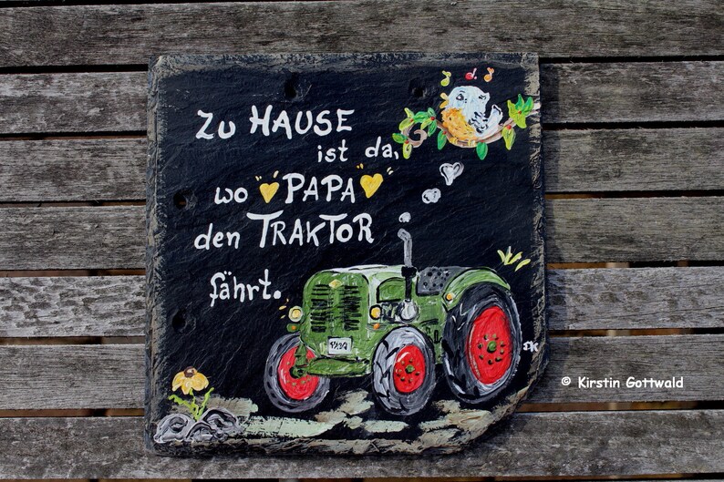 Puede incluir: Letrero de pizarra pintado a mano con un tractor verde y el texto alem&aacute;n "Zu Hause ist da, wo Papa den Traktor f&auml;hrt." Incluye detalles de p&aacute;jaros y flores. El letrero se muestra sobre una superficie de madera.