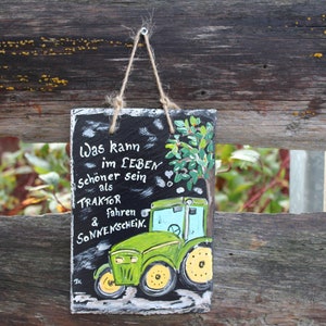 May include: A rectangular, hand-painted slate sign with a green tractor illustration and German text. The tractor is green with yellow wheels and a blue cab. The sign is suspended by a twine loop against a weathered wooden fence.