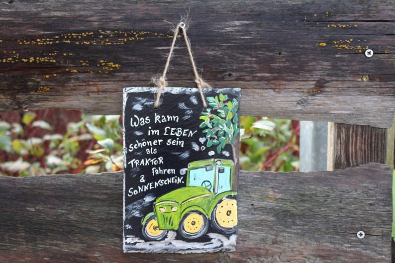 May include: A rectangular, hand-painted slate sign with a green tractor illustration and German text. The tractor is green with yellow wheels and a blue cab. The sign is suspended by a twine loop against a weathered wooden fence.
