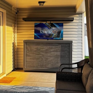 May include: A flat screen television displaying the Eyewitness News 9 logo mounted above a rectangular grey wooden cabinet with horizontal slats. The cabinet is positioned on a covered porch with light-coloured siding and a dark wicker chair.