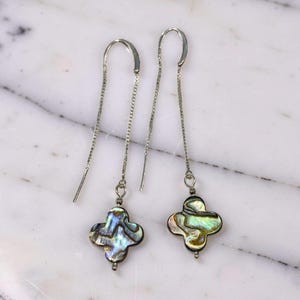May include: A pair of silver threader earrings. Each earring has a four-leaf clover-shaped pendant made of iridescent abalone shell. The earrings feature a delicate chain and a curved hook for easy wear.