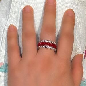 May include: A set of three stacking rings with red and silver beads. The rings are worn on a finger.
