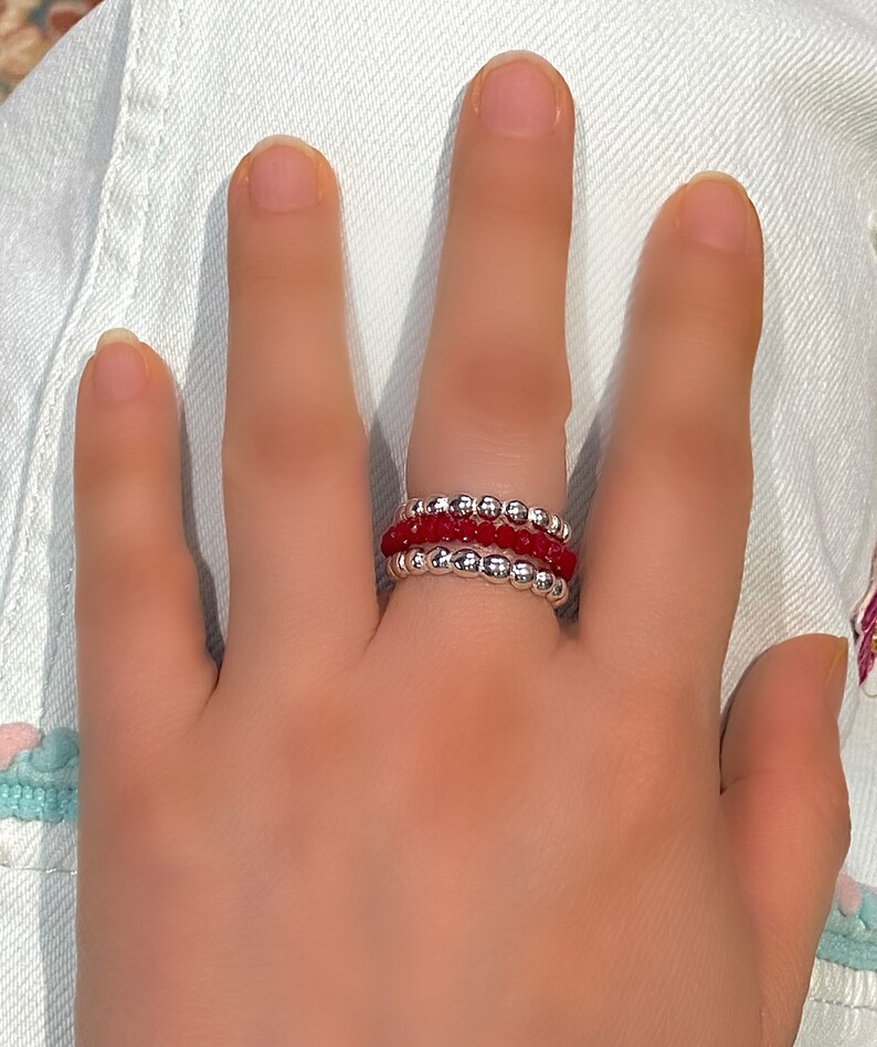 May include: A set of three stacking rings with red and silver beads. The rings are worn on a finger.