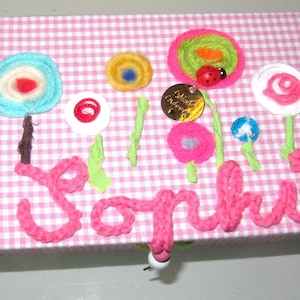 Könnte beinhalten: Handgefertigte Box aus rosa-weißem Vichy-Stoff, bestickt mit dem Namen "Sophie" in rosa Garn. Das Design umfasst bunte Garnblumen und einen Marienkäfer. Ein bezauberndes, personalisiertes Geschenk.