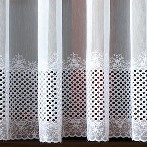 Può includere: Tende bianche trasparenti con un bordo decorativo in pizzo floreale e un motivo a pois. Le tende sono appese davanti a una finestra, permettendo alla luce di filtrare. Il design è elegante e adatto a un soggiorno o una camera da letto.