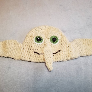 May include: A crocheted hat shaped like a Dobby the house-elf from the Harry Potter series. The hat is cream colored with green eyes and a long nose.