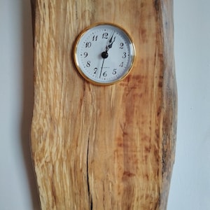 May include: A round, white clock face with black numbers and hands mounted on a rustic, wooden slab. The clock face has a gold rim and the words "QUARTZ" are printed on the face.