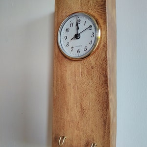 May include: A wooden key holder with a clock and two hooks. The clock has a white face with black numbers and hands. The wood is a light brown colour.