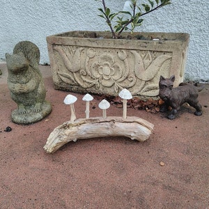 Puede incluir: Una rama de madera flotante decorativa con cinco setas de cerámica blanca unidas a ella. La rama está sobre una superficie de grava roja. Hay una pequeña estatua de ardilla y una pequeña estatua de perro a cada lado de la rama.