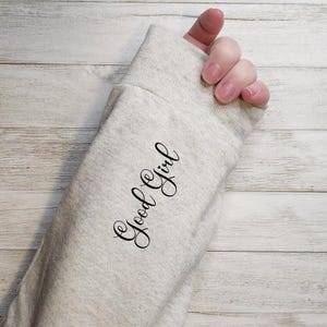 Op de afbeelding: Een lichtgrijze sweatshirtmouw met de cursieve tekst "Good Girl" in het zwart gedrukt. De mouw is bij de pols afgezet. De achtergrond is een lichte houtnerf.