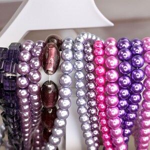 May include: A selection of beaded necklaces in shades of purple, pink, and silver. The beads vary in size and shape, including round pearls and square-cut beads. The necklaces are displayed on a white rack.