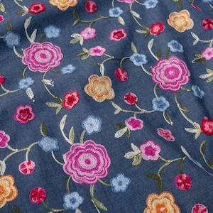 Könnte beinhalten: Nahaufnahme eines Jeansstoffs mit gestickten Blumenmustern. Die Stickerei zeigt Blumen in Rosa, Rot, Gelb und Blau, mit grünen Stielen und Blättern. Der Stoff hat eine leicht zerknitterte Textur.