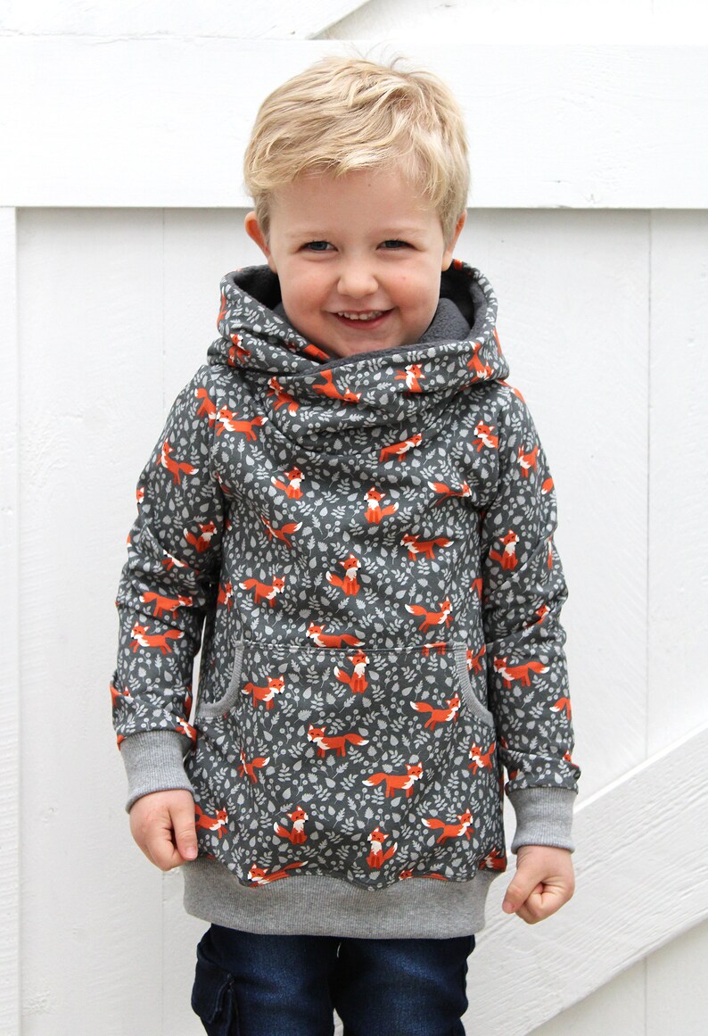 K&ouml;nnte beinhalten: Ein kleines Kind tr&auml;gt einen grauen Kapuzenpullover mit einem Fuchs-Druck. Der Pullover hat eine gro&szlig;e Kapuze und eine K&auml;ngurutasche. Das Kind l&auml;chelt und schaut in die Kamera.