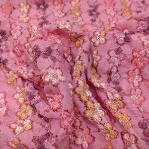 Pode incluir: Tecido de renda floral rosa com um padrão delicado de flores em tons de rosa, amarelo e branco. O tecido tem uma textura macia e transparente e um brilho sutil.