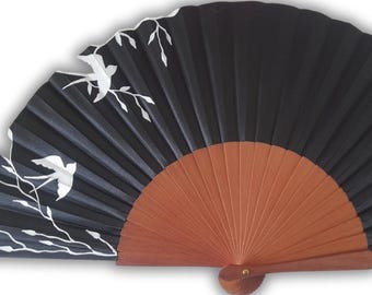 May include: A black hand fan with a wooden handle and a semi-circular shape. The fan features white bird and branch designs. The fan is made of pleated material and has a wooden handle. The fan is a decorative accessory.